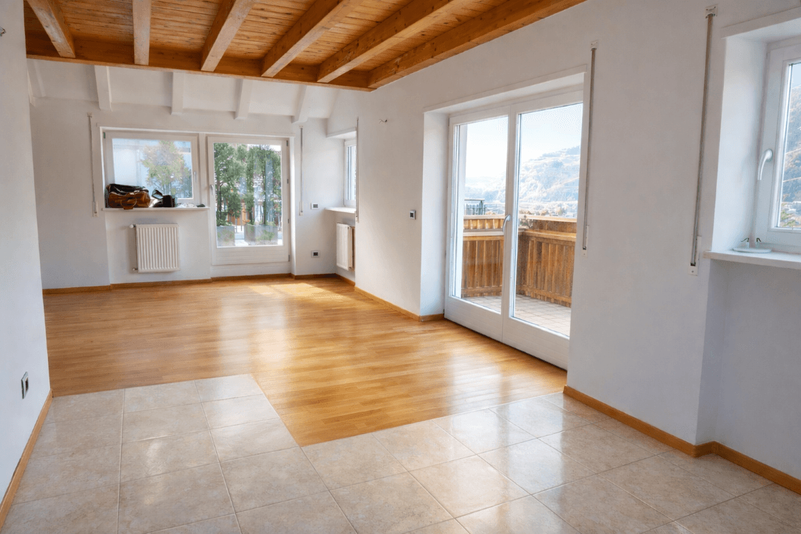 Heller, leerer Raum mit gemischtem Bodenbelag, Holzbalkendecke und Zugang zum Balkon.