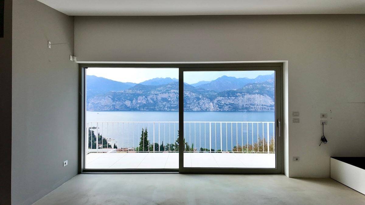 Heller Raum mit Betonboden und großen Glasschiebetüren, die einen weiten Blick auf den See und die Berge freigeben.