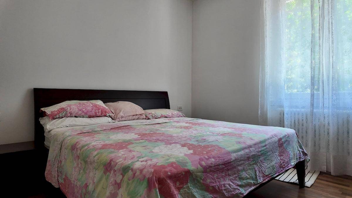 Schlafzimmer mit Doppelbett und floraler Bettwäsche, hell durch großes Fenster mit Vorhängen.