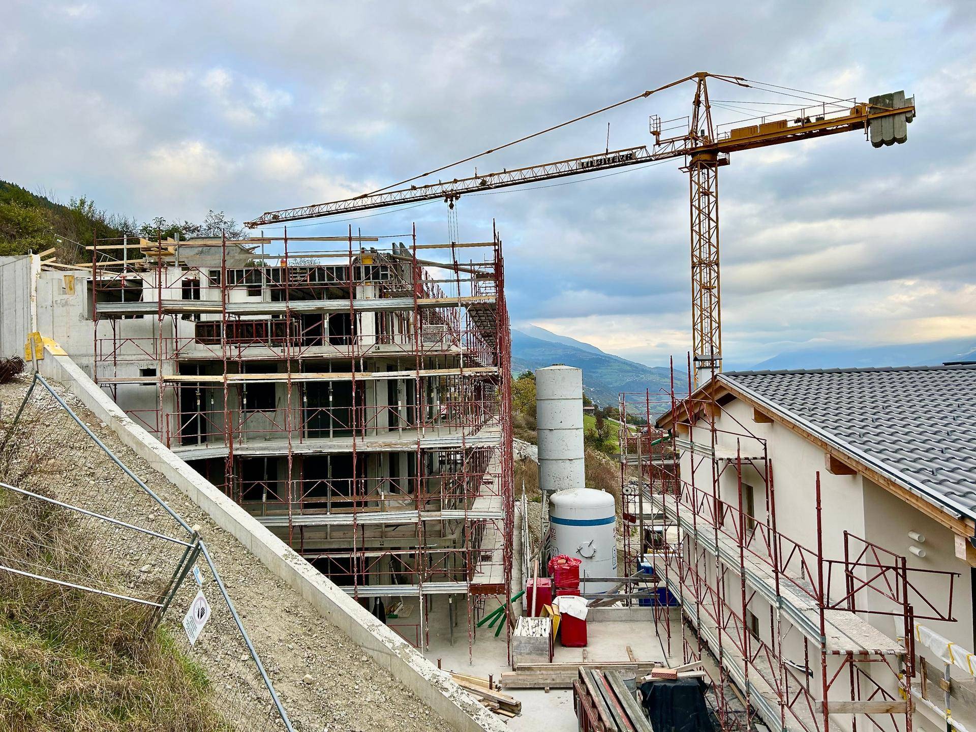 Baustelle mit mehreren Gebäuden im Bau, umgeben von einer hügeligen Landschaft unter bewölktem Himmel.