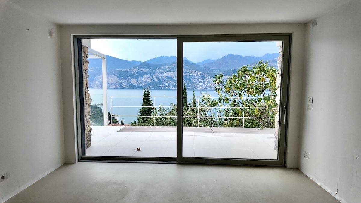 Leerer Raum mit Betonboden und großen Glasschiebetüren, die zu einer Terrasse mit Seeblick führen.