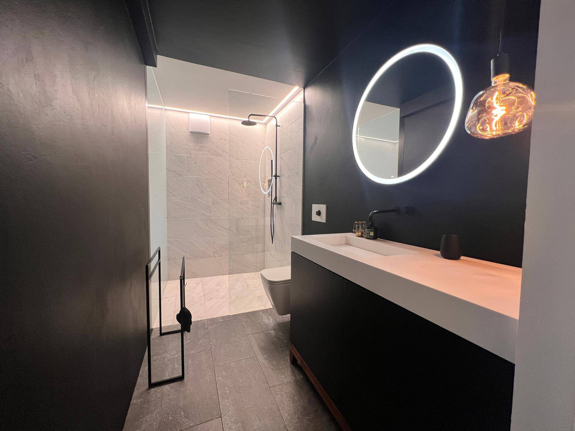 Elegantes Badezimmer mit dunklen Wänden, beleuchtetem Rundspiegel und großer Dusche.