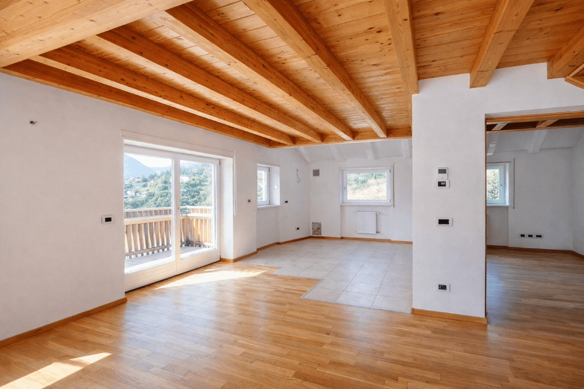 Heller, leerer Raum mit gemischtem Bodenbelag, Holzbalkendecke und Balkonzugang.