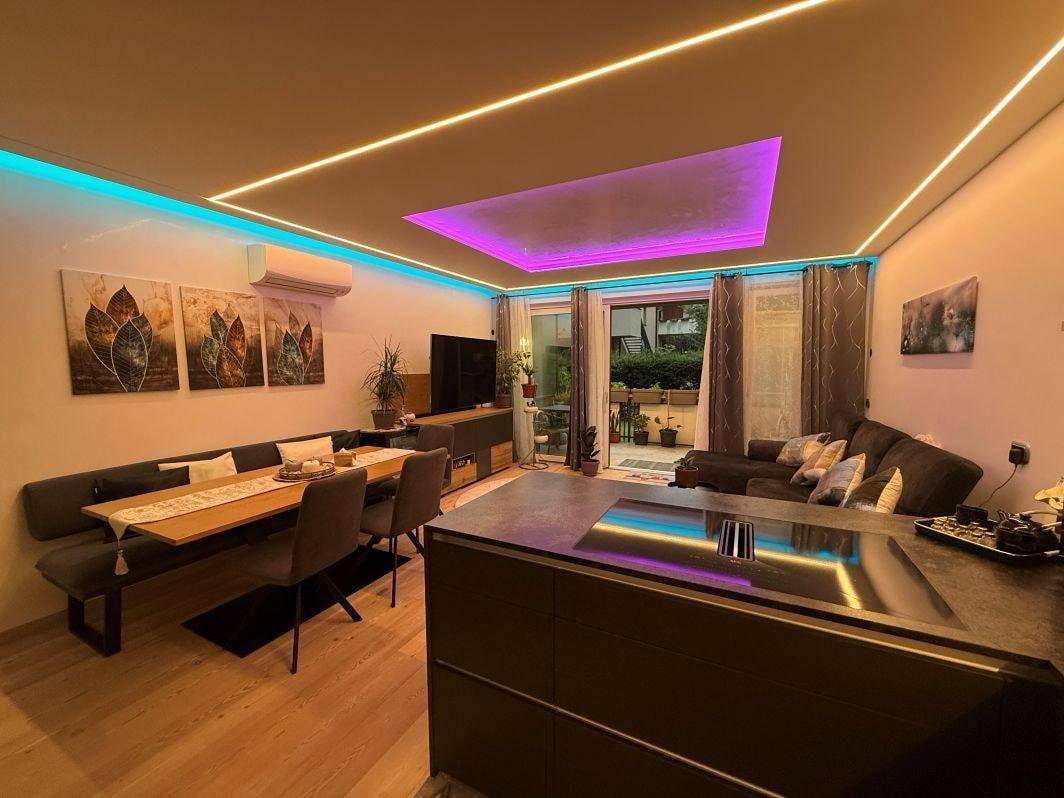 Offener Wohnbereich mit Esstisch, Sofa und integrierter Küche, beleuchtet mit farbiger LED-Beleuchtung.