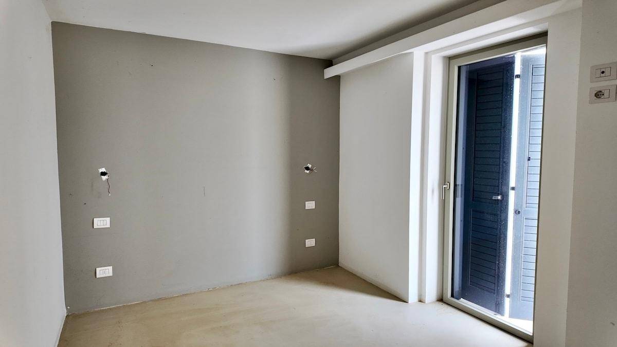 Leeres Zimmer mit Betonboden und grauen Wänden, mit einer Tür, die zu einem Außenbereich führt.