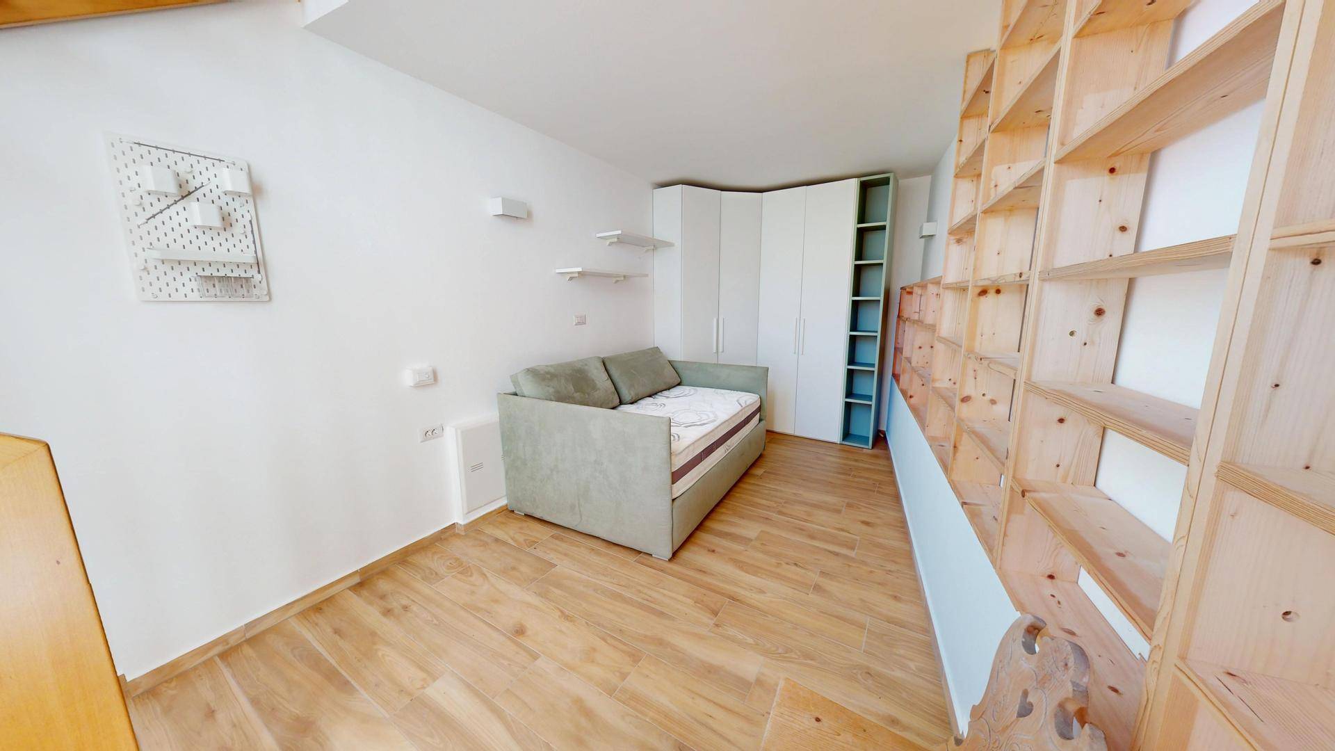 Funktionales Zimmer mit Schlafsofa, weißem Kleiderschrank und integrierten Holzregalen für Stauraum.