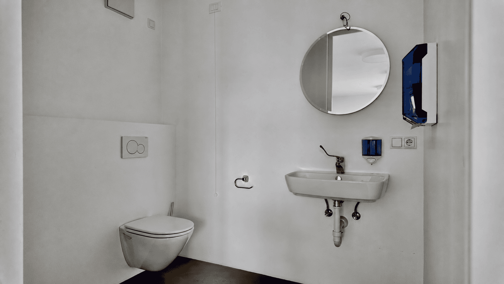 Modernes Badezimmer mit wandhängender Toilette, Waschbecken, rundem Spiegel und einem blauen Spender.