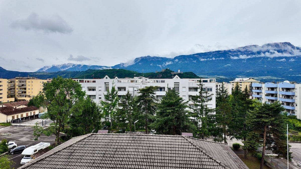 Blick auf ein Wohngebiet mit Gebäuden, Bäumen und Bergen unter bewölktem Himmel.