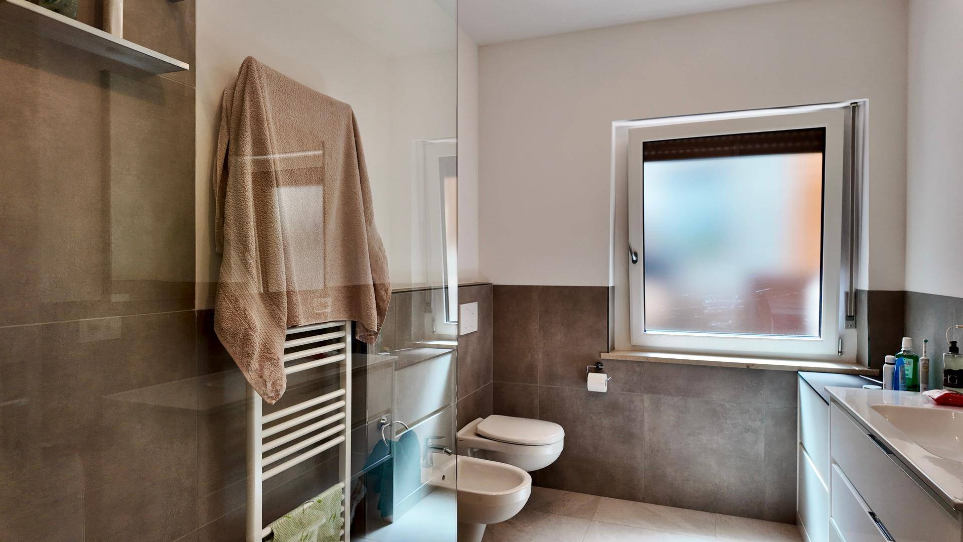 Modernes Badezimmer mit grauen Fliesen, WC, Bidet, Waschtisch und Fenster mit Milchglas.