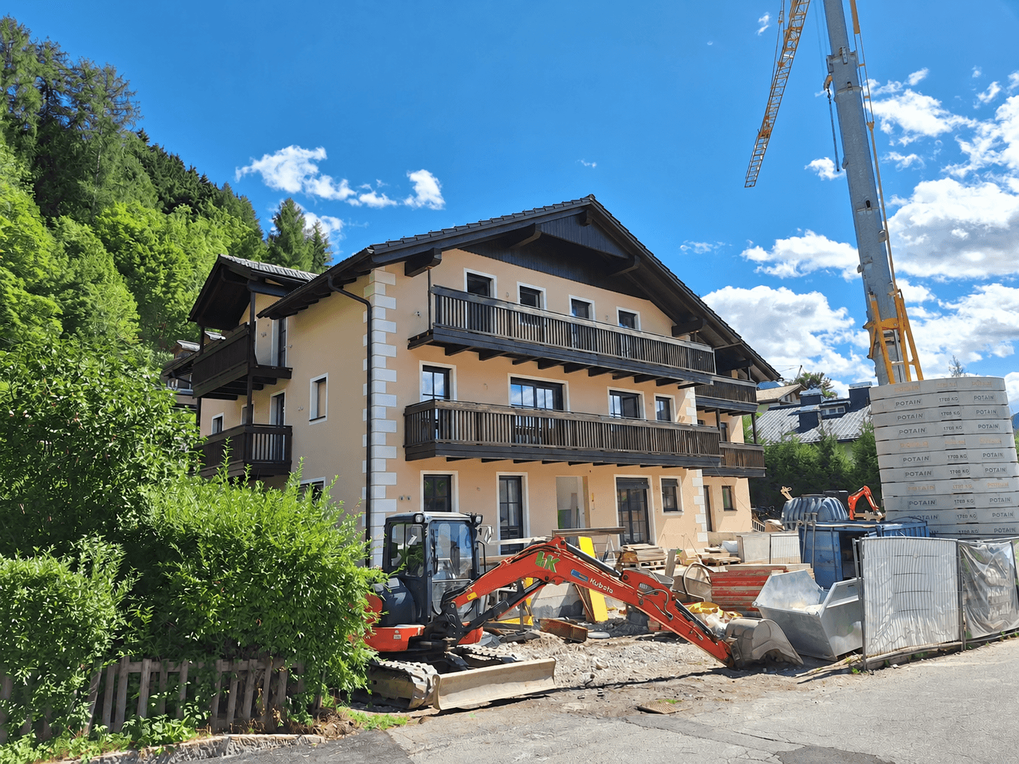 Neubau im alpinen Stil mit Balkonen, umgeben von Baustelle und grüner Berglandschaft.