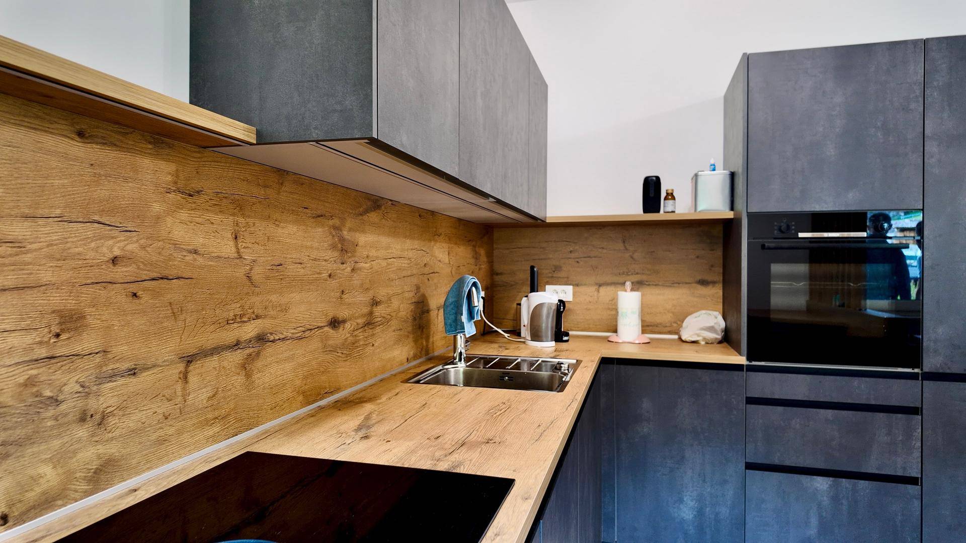 Moderne Küche mit Spülbecken, Induktionskochfeld, Backofen und dunklen Schränken mit Holzakzenten.