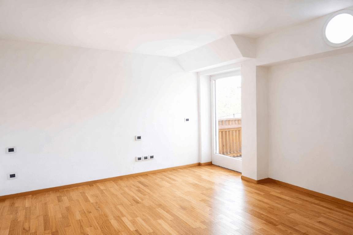 Leeres Schlafzimmer mit Holzboden, weißen Wänden und Balkonzugang.
