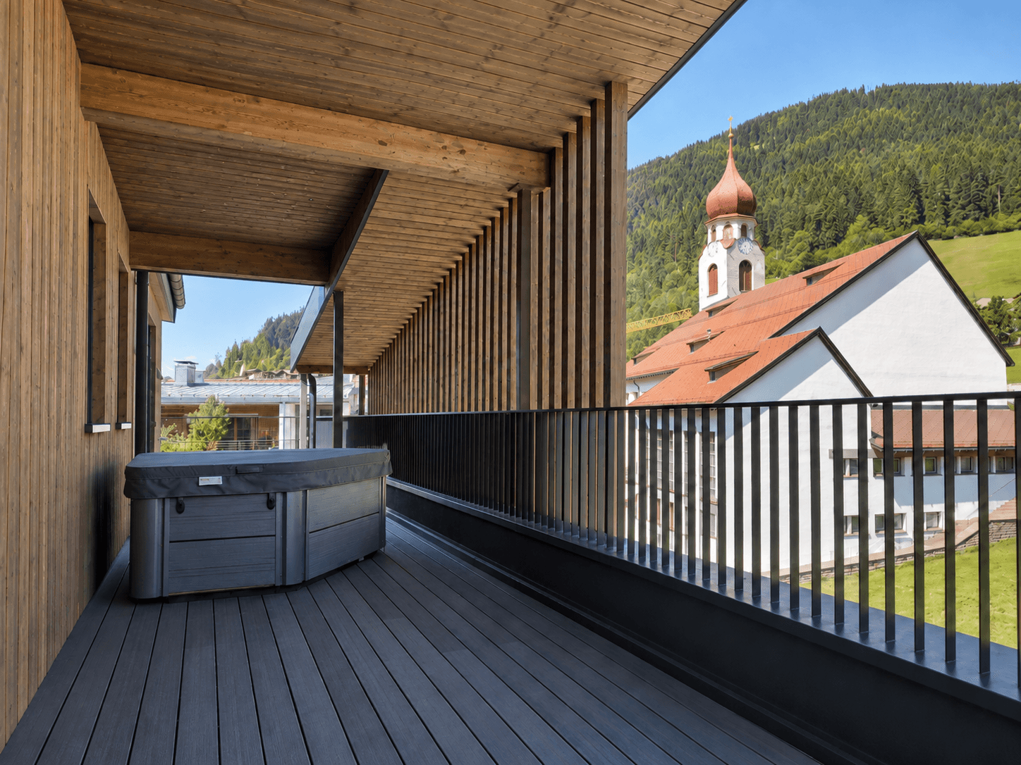 Überdachte Terrasse mit Whirlpool und Blick auf ein charmantes Dorf mit Kirchturm und grüne Hügel.