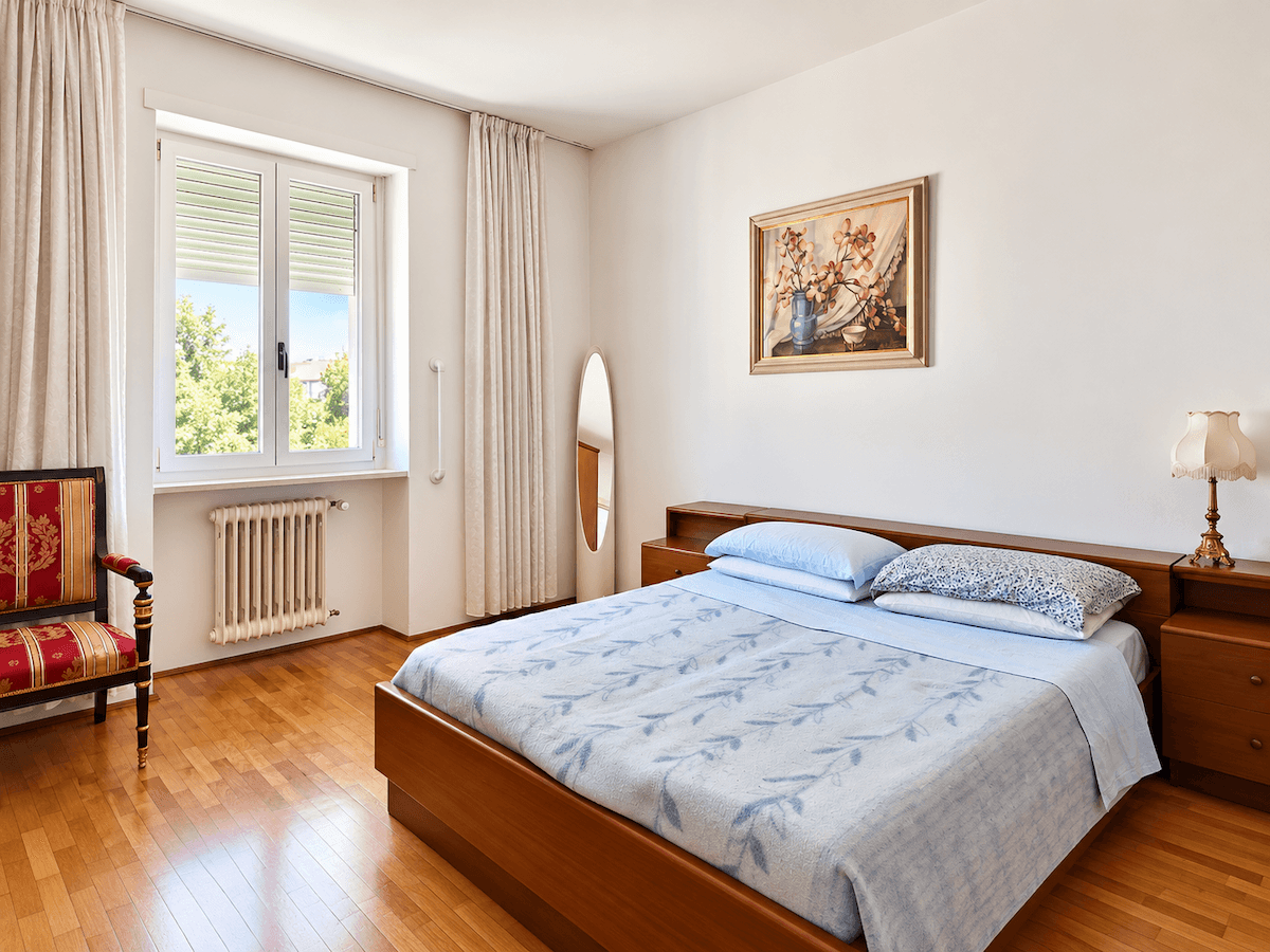Helles Schlafzimmer mit Doppelbett, Fenster mit Rollläden und einem klassischen Heizkörper.