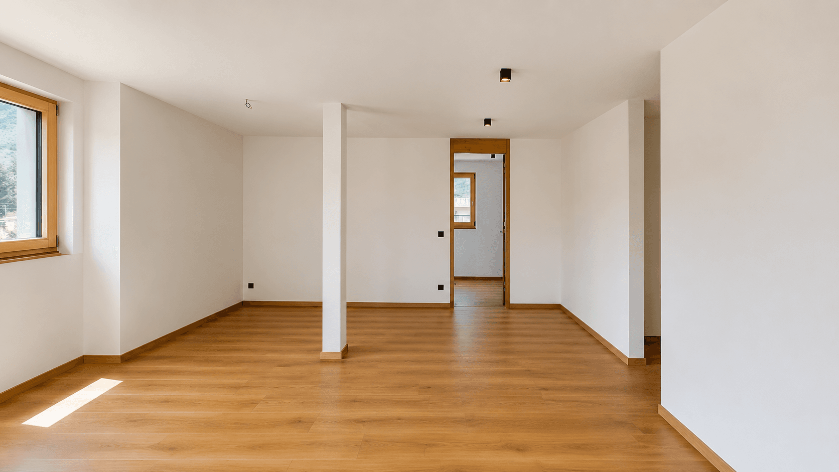 Heller Raum mit Holzboden und Fenster, der in einen weiteren Bereich führt.