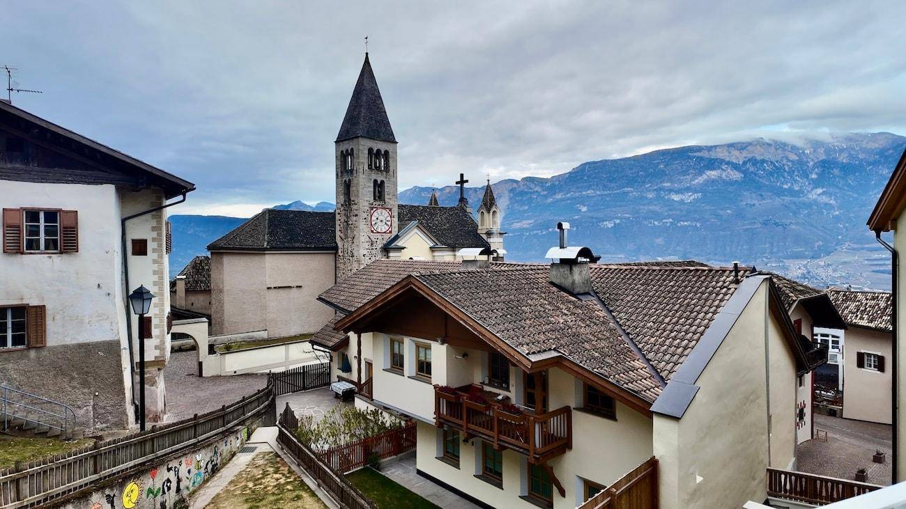 Panoramablick auf ein malerisches Dorf mit Kirchturm und majestätischen Bergen im Hintergrund.