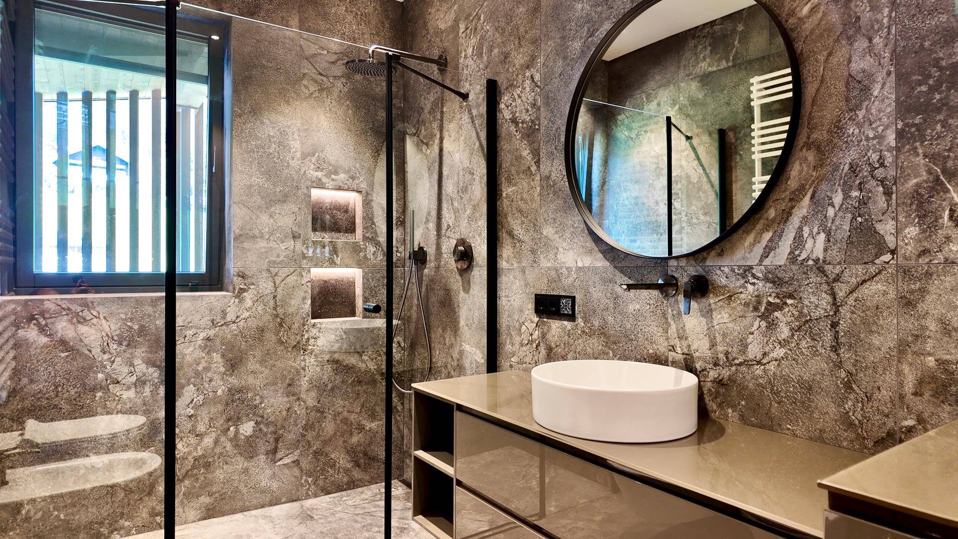 Modernes Badezimmer mit dunklen Fliesen, rundem Spiegel, Einzelwaschbecken und begehbarer Regendusche.