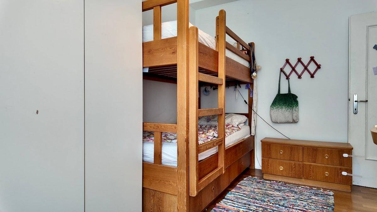 Kinderzimmer mit Etagenbett, Kommode und buntem Teppich, hell durch das Fenster.