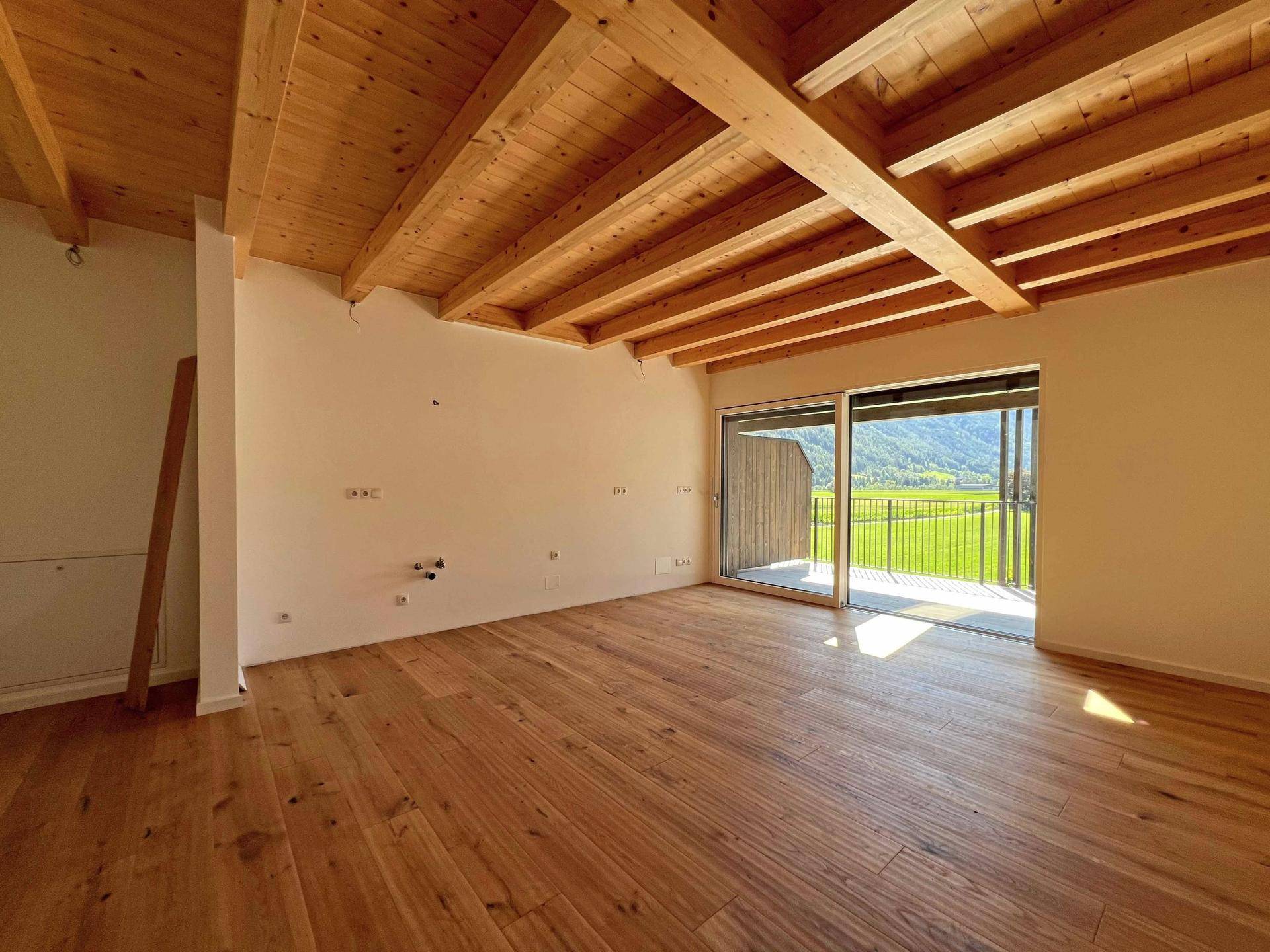 Geräumiger Wohnbereich mit Holzboden, sichtbaren Deckenbalken und großem Fenster mit Balkonzugang.