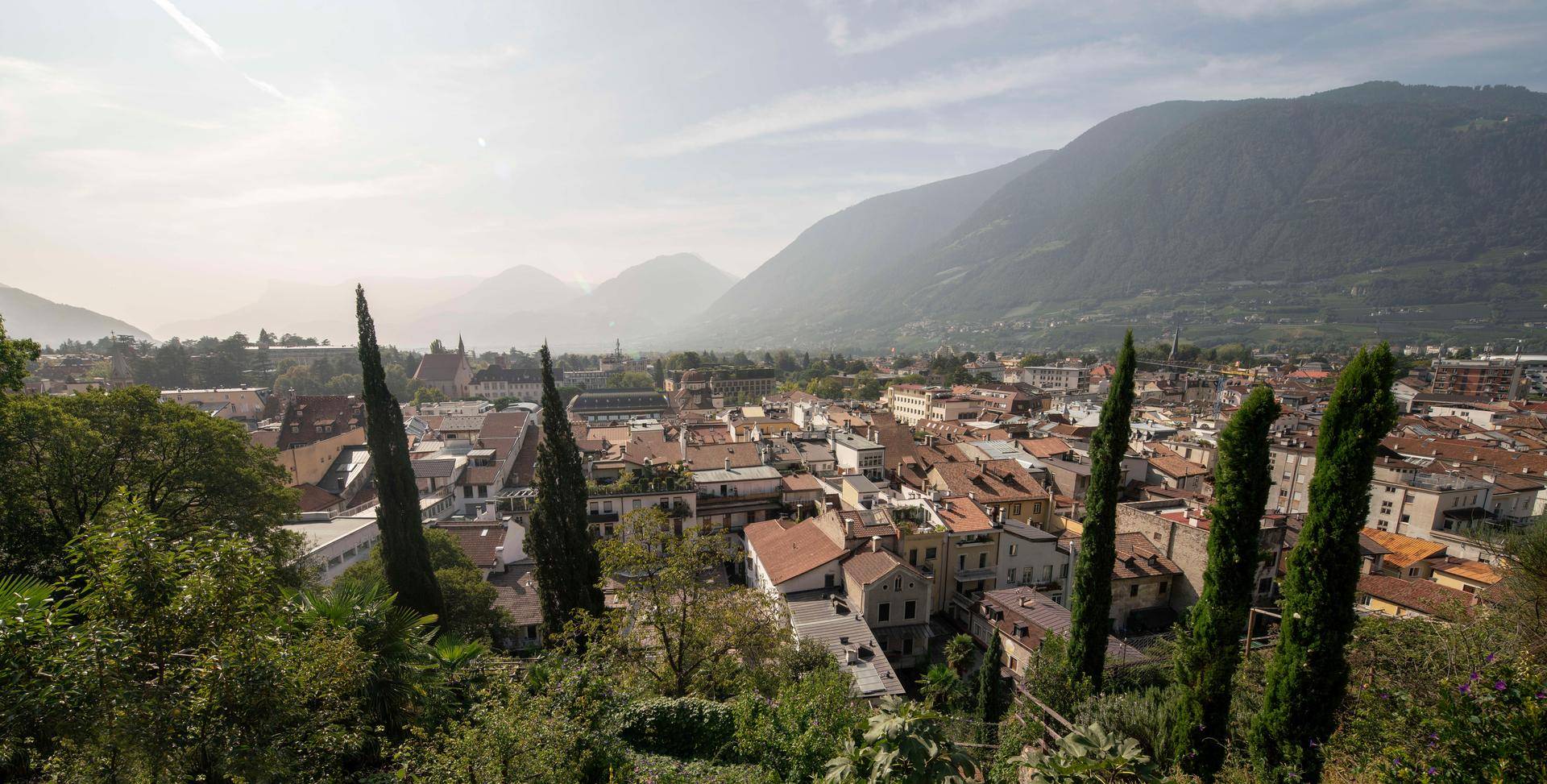 Weitläufiger Blick auf eine malerische Stadt im Tal, umgeben von Bergen und hohen Bäumen.