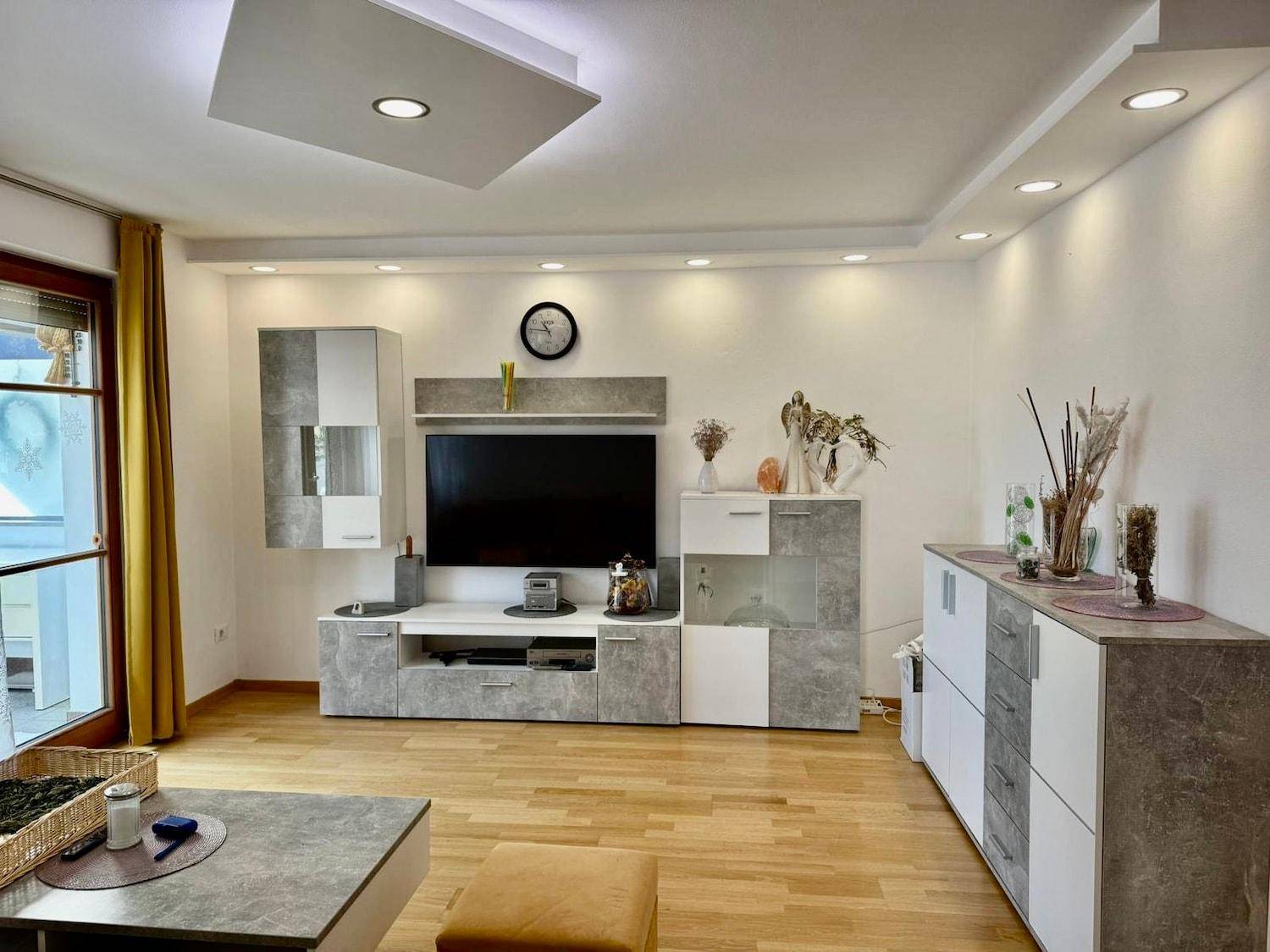 Modern eingerichtetes Wohnzimmer mit TV-Wand, indirekter Beleuchtung und hellem Parkettboden.