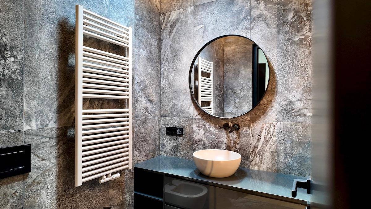 Modernes Badezimmer mit grauen Fliesen, rundem Spiegel, Aufsatzwaschbecken und Handtuchheizkörper.