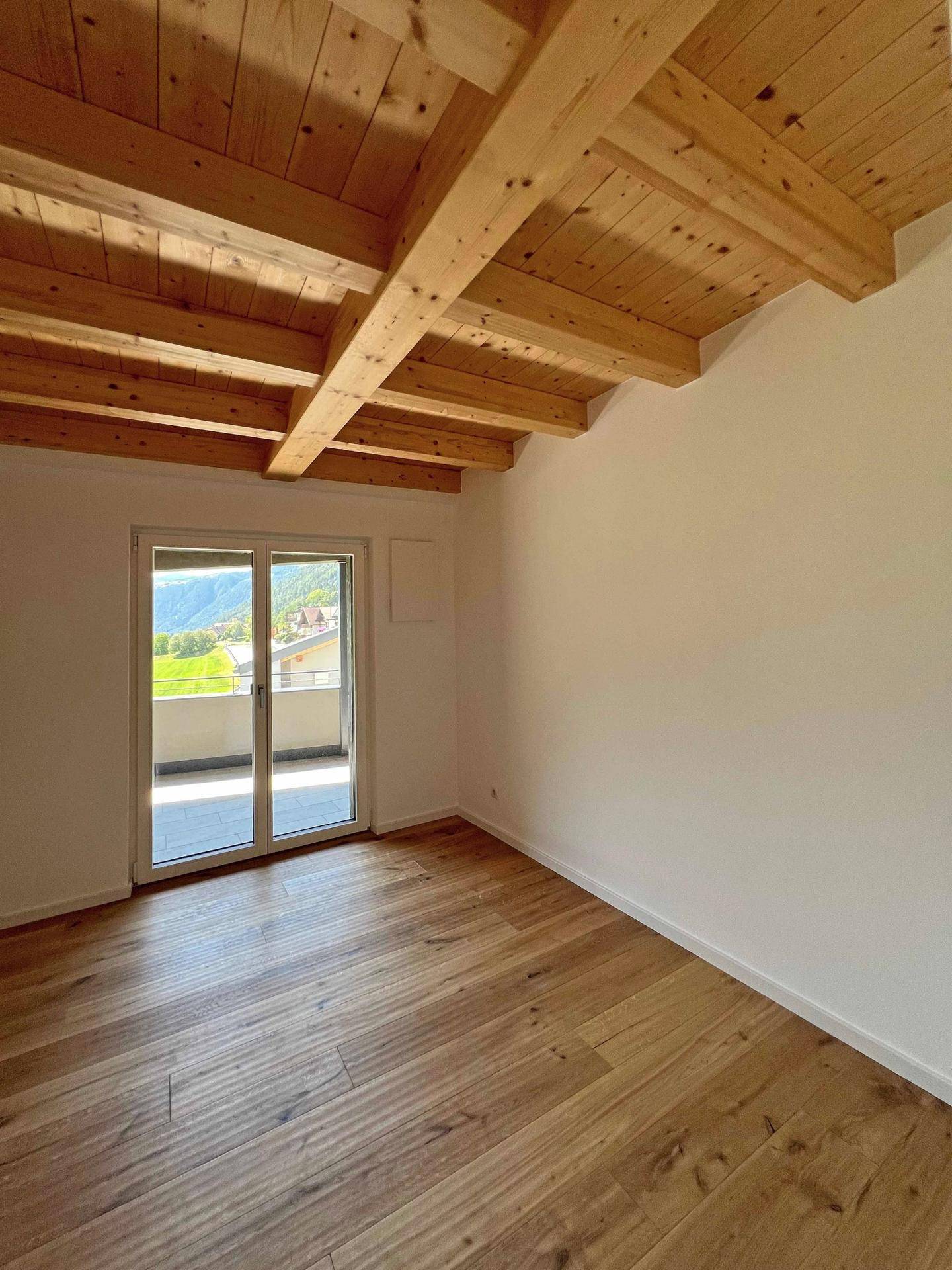 Helles Zimmer mit Holzboden, Holzbalkendecke und Zugang zu einem Balkon mit Blick ins Grüne.