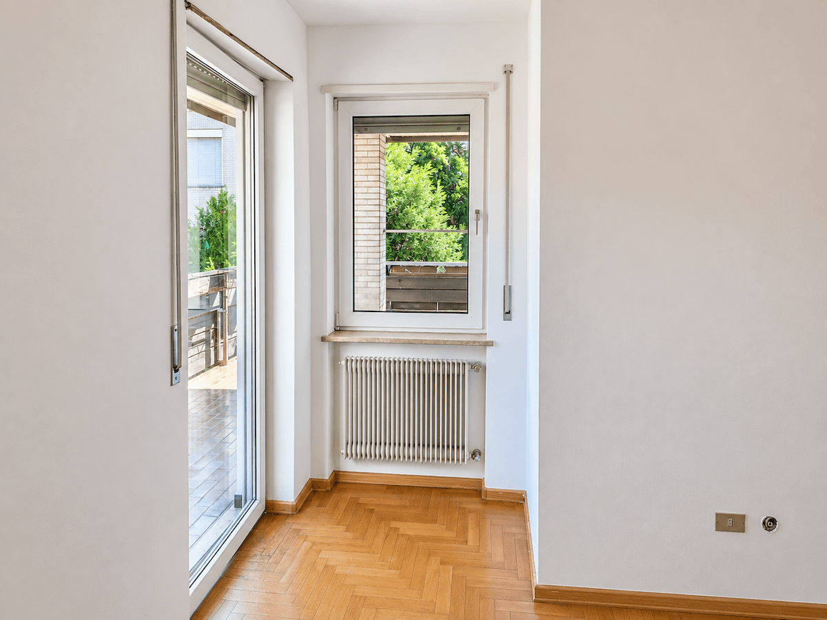 Helles Zimmer mit Fischgrätparkett, Heizkörper und Zugang zu einem Balkon.