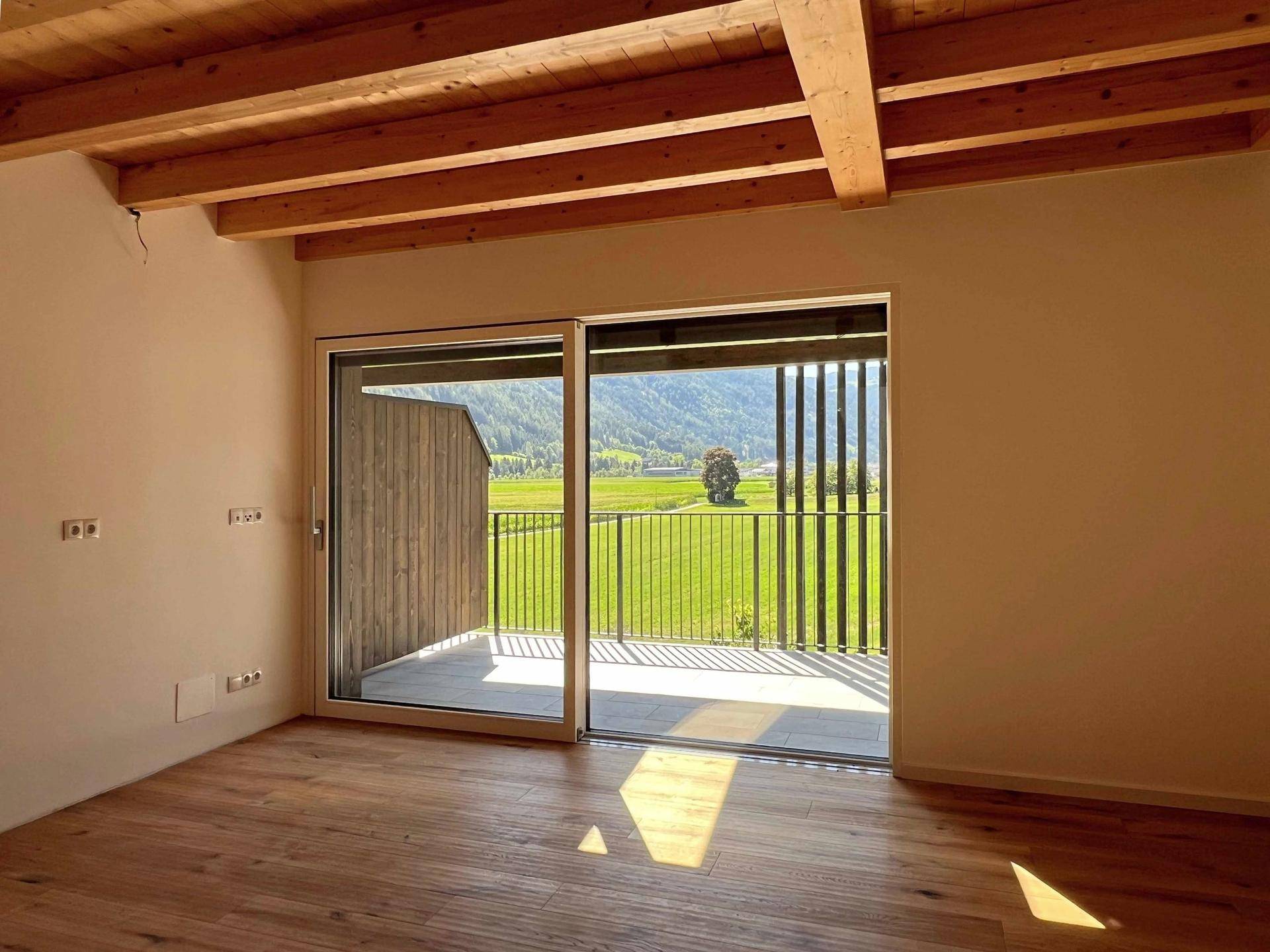 Schlafzimmer mit Holzboden, sichtbaren Deckenbalken und großem Schiebefenster zum Balkon mit Aussicht.
