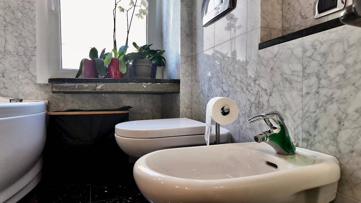 Badezimmer mit Bidet, Toilette und Fenster, umgeben von Marmorfliesen.