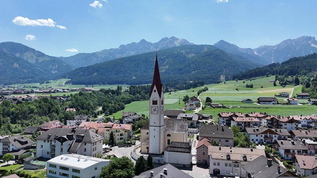 Panoramablick auf ein malerisches Dorf mit Kirche, umgeben von grünen Hügeln und Bergen.