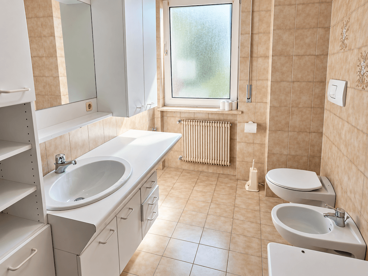 Badezimmer mit beigefarbenen Fliesen, Einzelwaschbecken, Toilette, Bidet und Fenster.