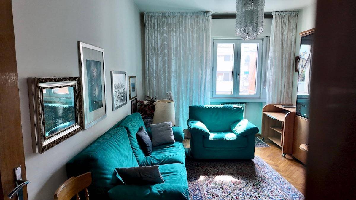Wohnzimmer mit türkisfarbenem Sofa, Teppich und einem Fenster mit Vorhängen.