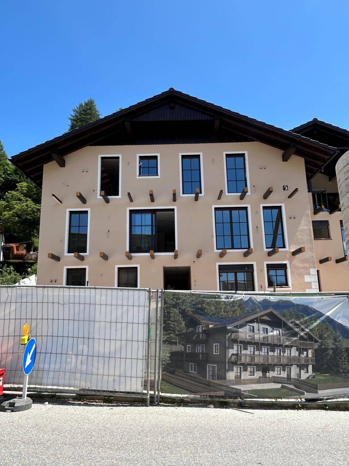 Vorderansicht des Neubaus von der Straße aus, mit Bauzaun und Visualisierung des fertigen Gebäudes.