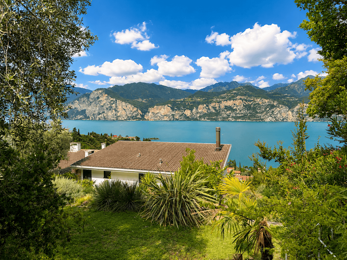 Haus mit braunem Dach, umgeben von üppigem Grün, mit weitem Blick auf den See und die Berge.