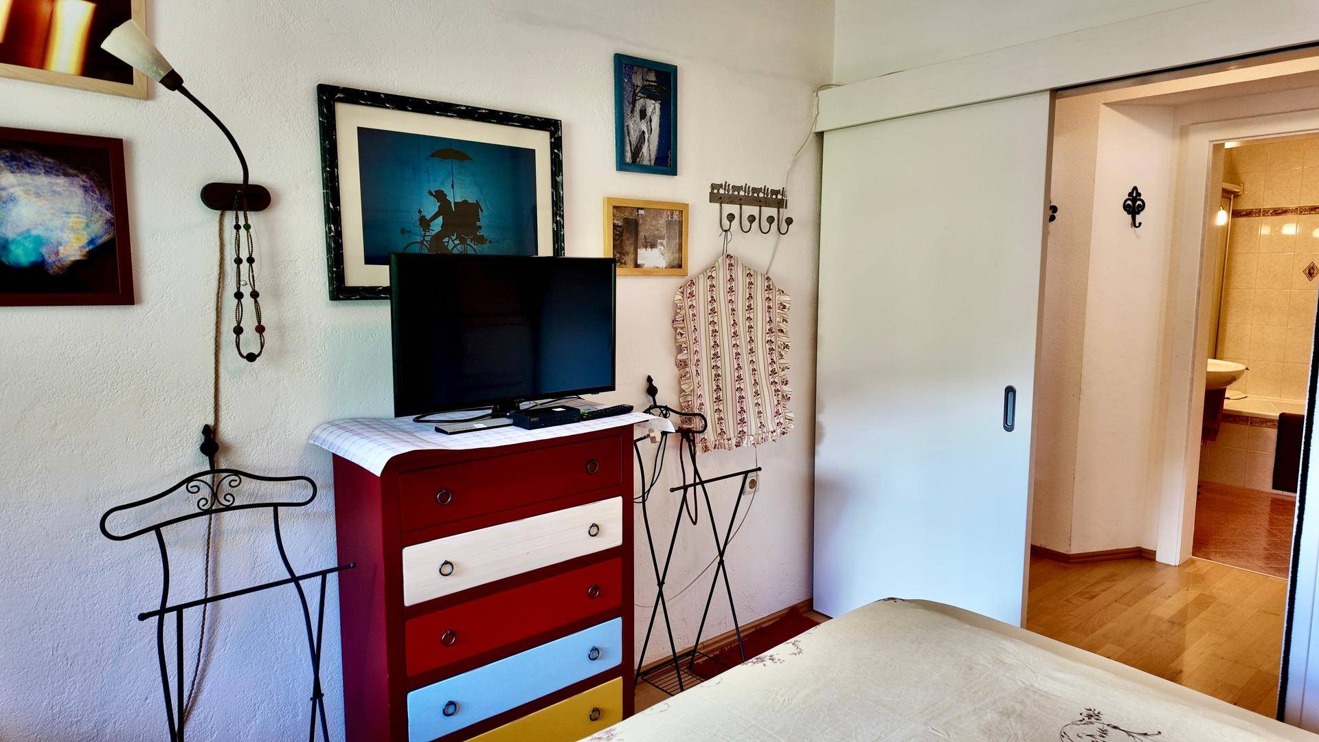 Schlafzimmer mit farbenfroher Kommode, Fernseher und Schiebetür zu einem angrenzenden Badezimmer.
