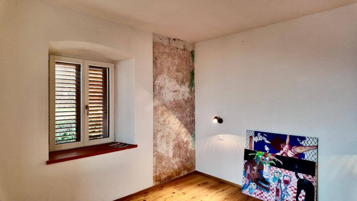 Heller Raum mit Holzboden, Fenster mit Jalousien und einer Wand mit Putzstruktur.