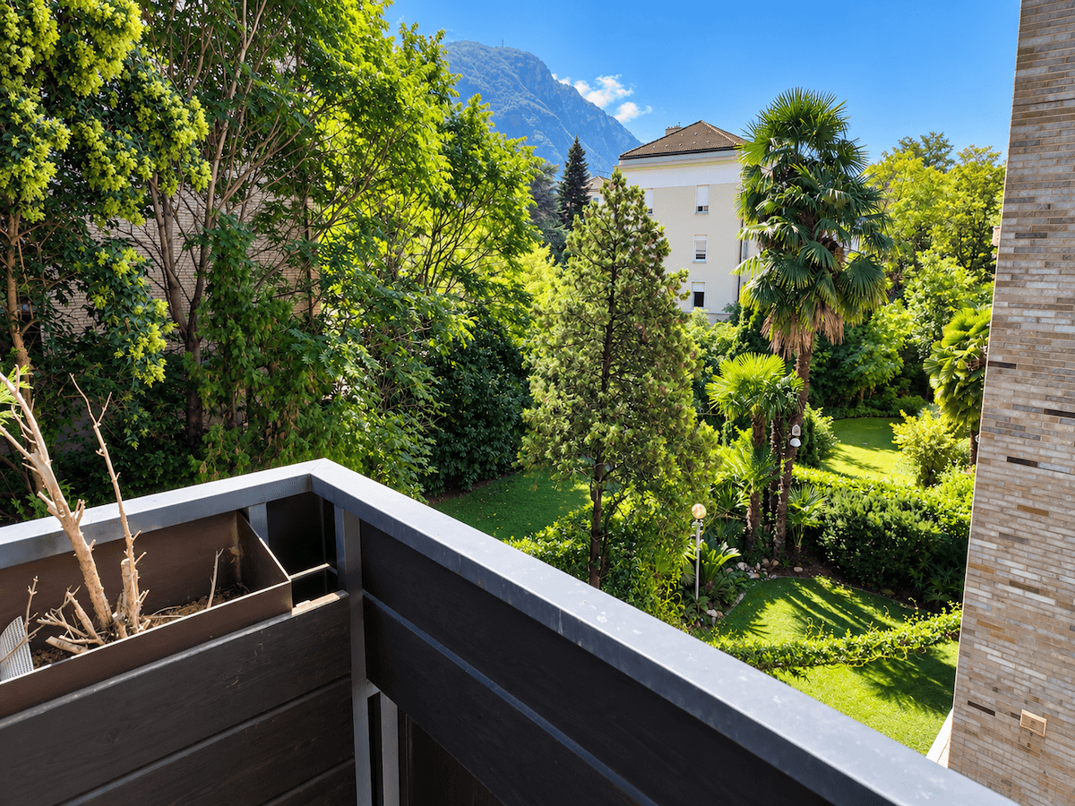 Blick von einem Balkon auf einen üppigen grünen Garten mit Bäumen und Bergen im Hintergrund.