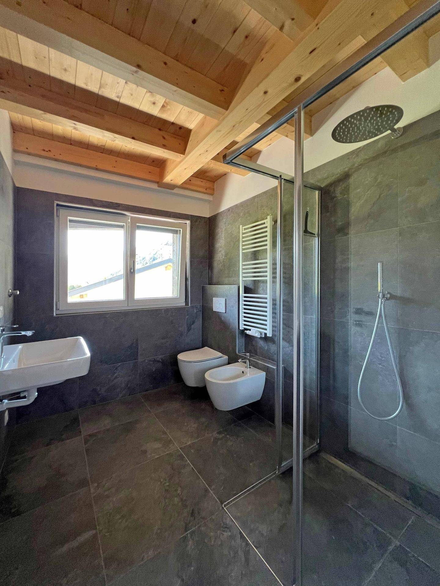 Modernes Badezimmer mit dunklen Fliesen, Holzbalkendecke, Fenster und einer geräumigen Regendusche.