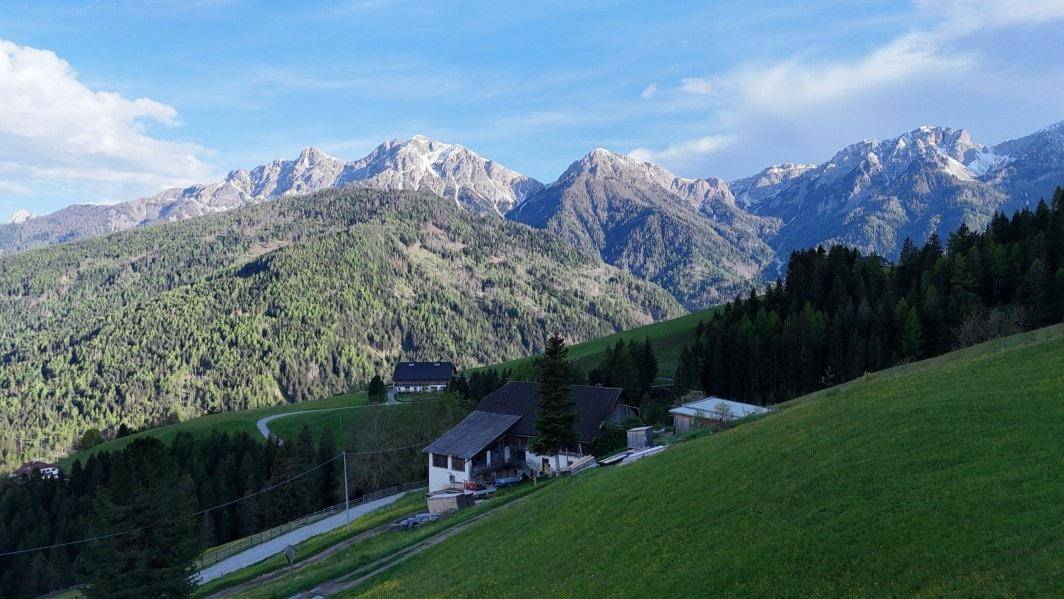 Traditionelles Haus in alpiner Umgebung, umgeben von grünen Wiesen und einer beeindruckenden Bergkette.