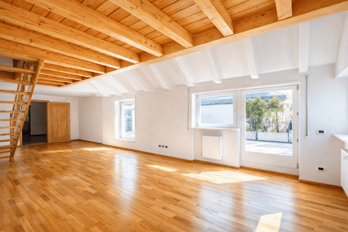 Geräumiger, leerer Raum mit Holzboden, sichtbaren Deckenbalken und Holztreppe.