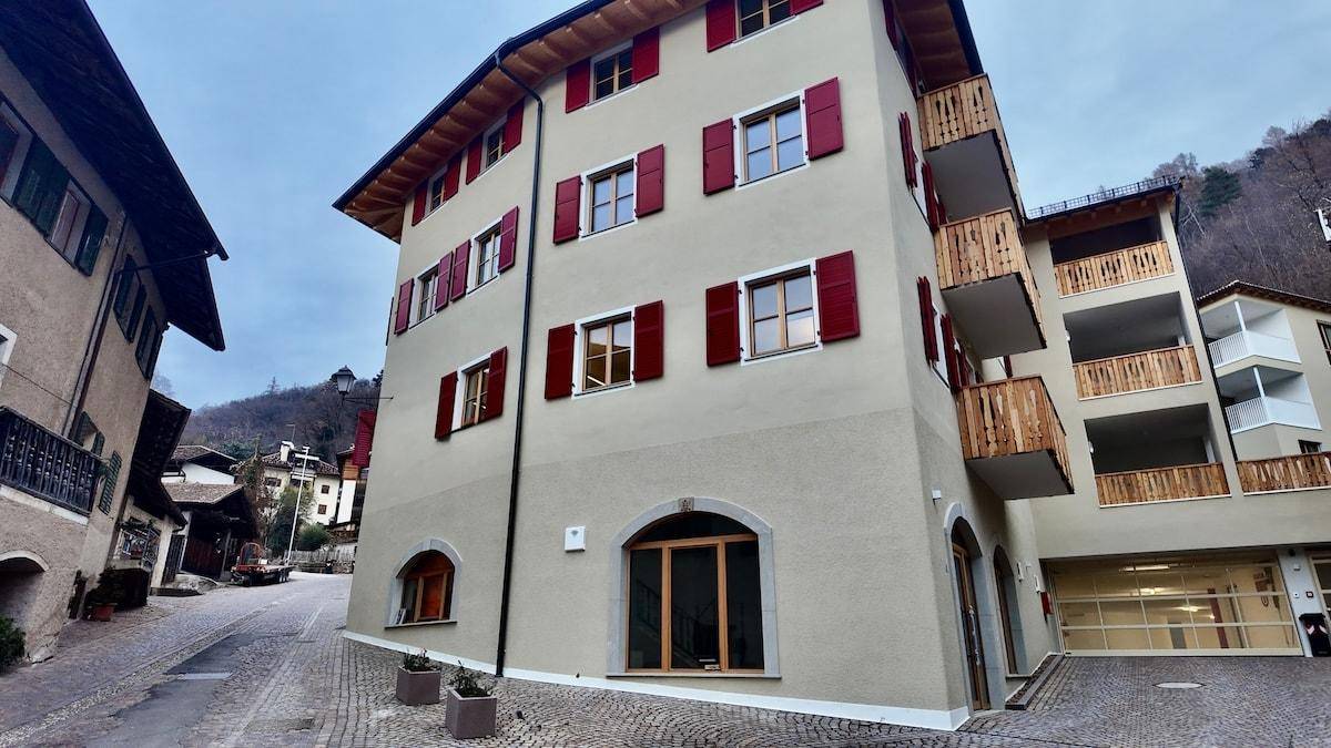 Gesamtansicht eines modernen Gebäudes mit Holzbalkonen, roten Fensterläden und gepflasterter Straße.