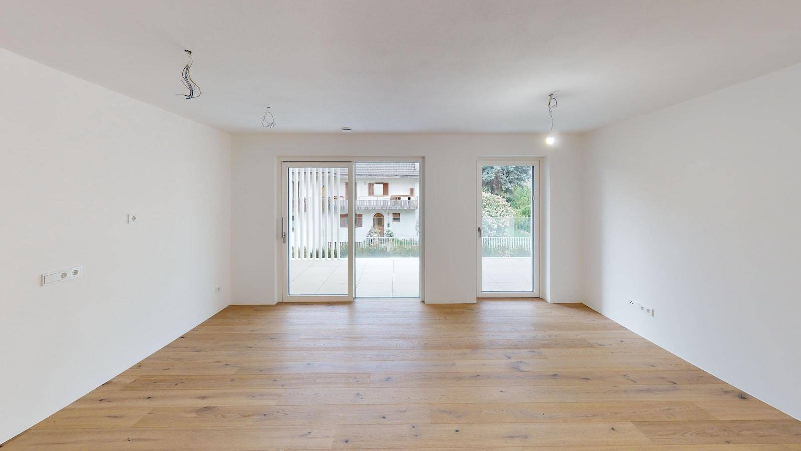 Großer, heller Raum mit Holzboden und zwei Fenstertüren, die zu einem Außenbereich führen.