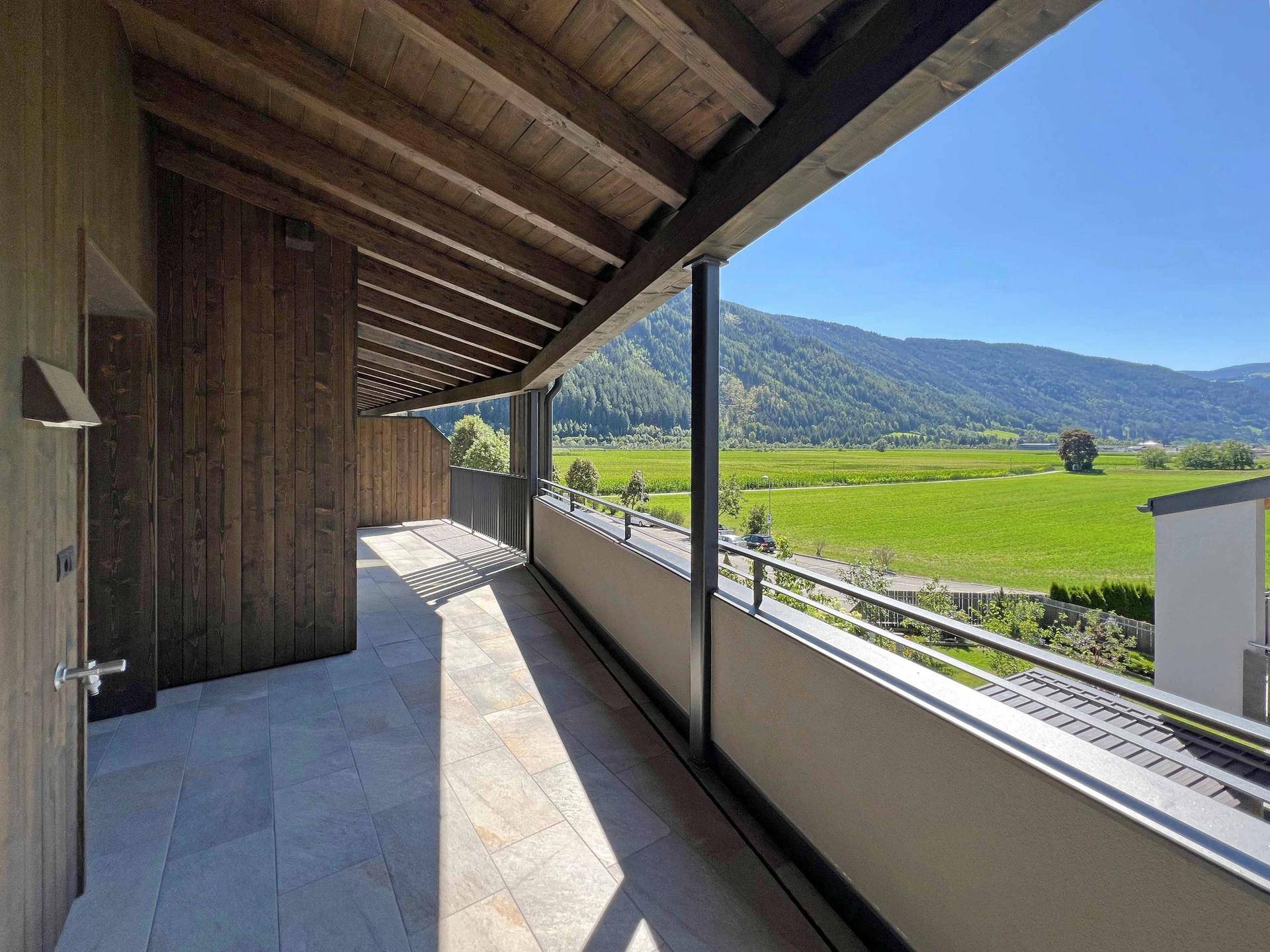 Lange Terrasse mit Holzwänden und -decke, gefliestem Boden und Panoramablick auf die umliegende Natur.