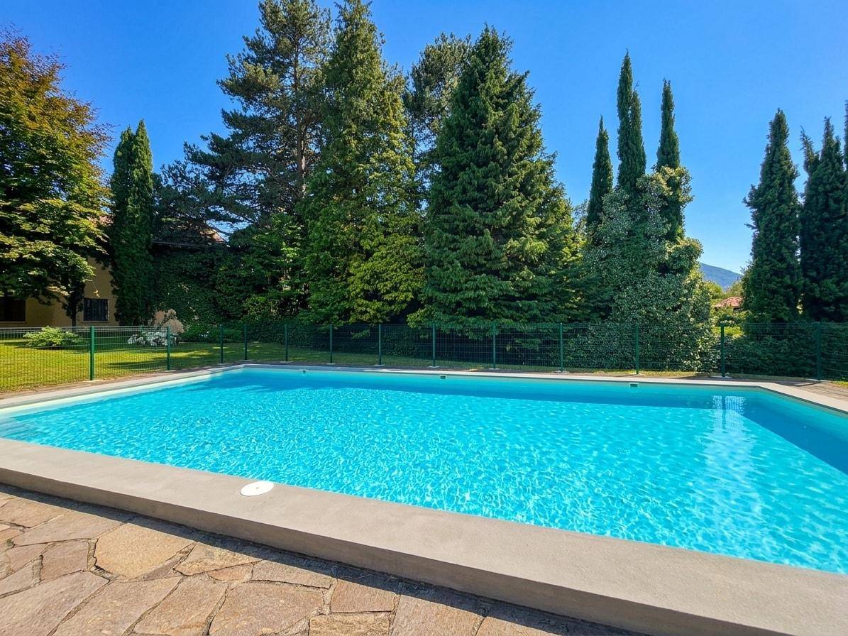 Weitläufiger Außenpoolbereich mit viel Grün und klarem Wasser unter blauem Himmel.