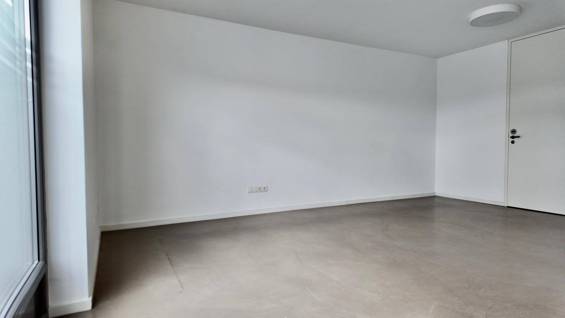 Heller Innenraum mit einer großen Fensterfront, die viel Tageslicht hereinlässt, und einem schlichten Boden.
