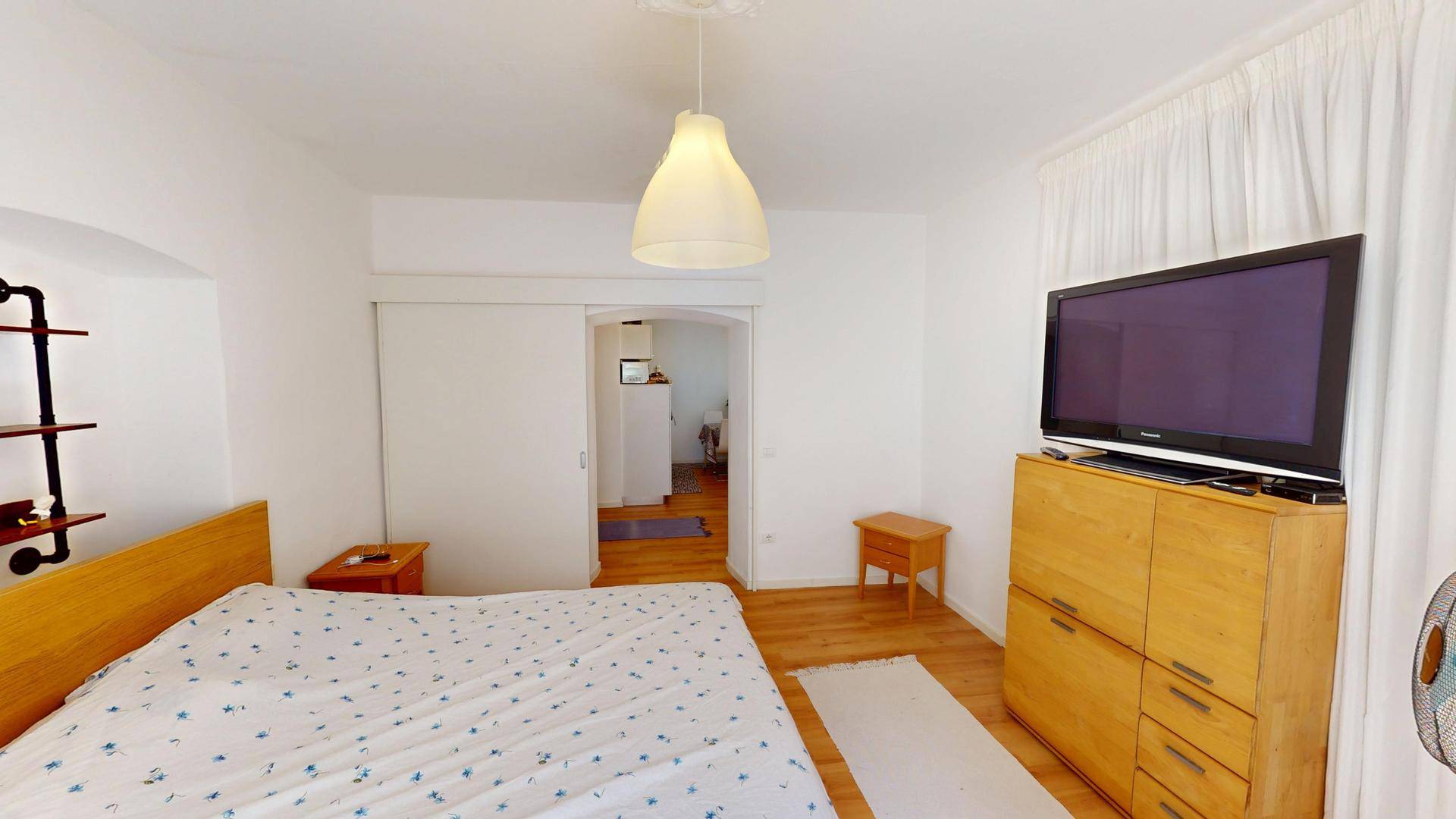 Helles Schlafzimmer mit Holzboden, Fernseher und Zugang zu einem weiteren Raum durch eine Schiebetür.
