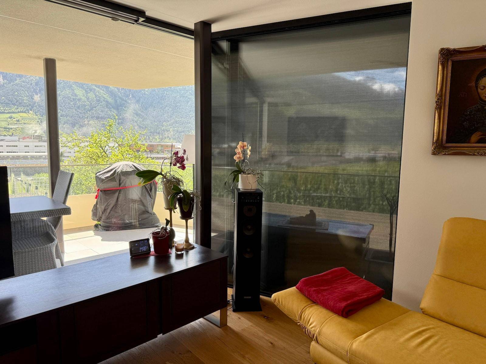 Gemütliches Wohnzimmer mit gelbem Sofa, Fernseher und Panoramablick auf Weinberge und Berge.