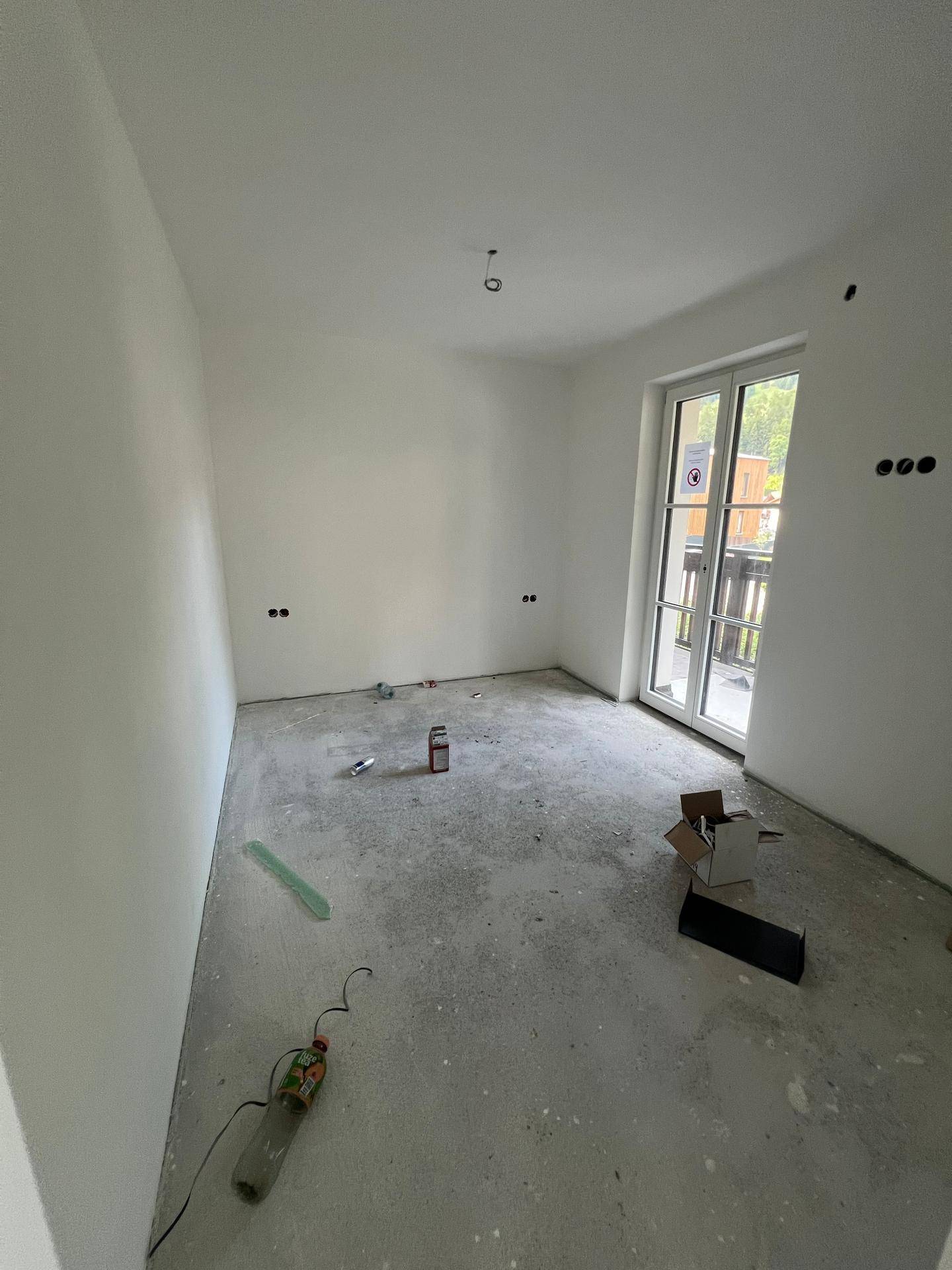 Unfertiges Schlafzimmer mit weiß gestrichenen Wänden, Betonboden und Balkonzugang.