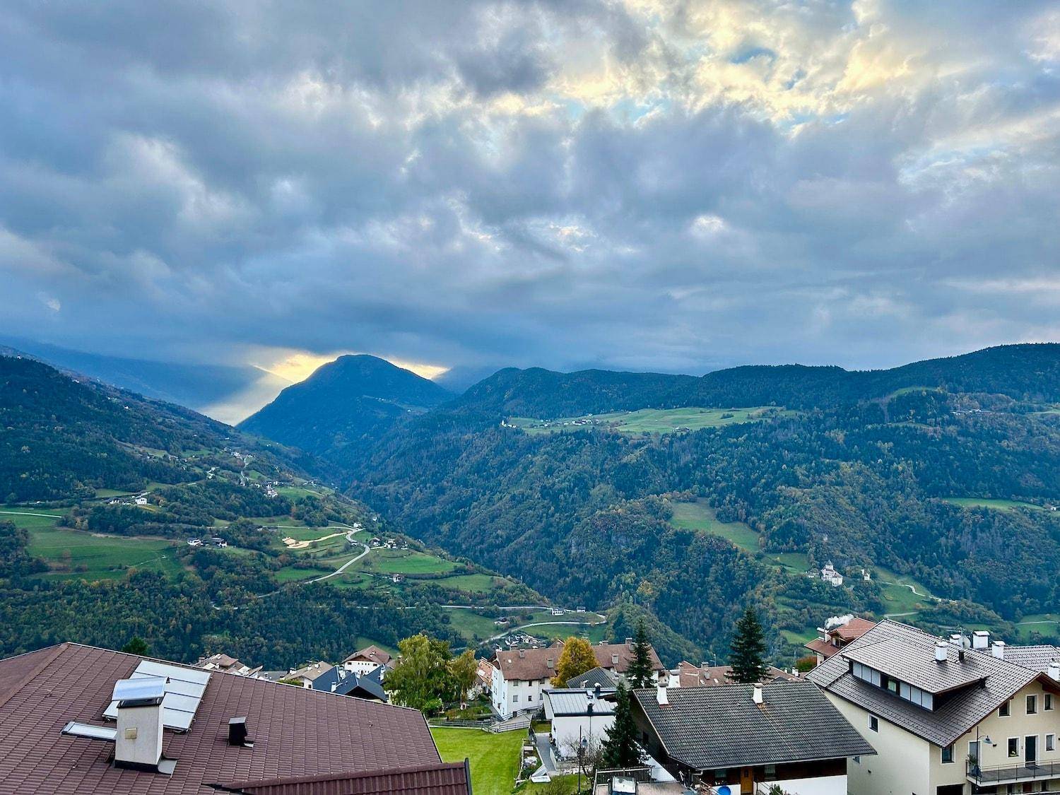 Malerische Berglandschaft mit bewaldeten Hängen und verstreuten Häusern unter bewölktem Himmel.